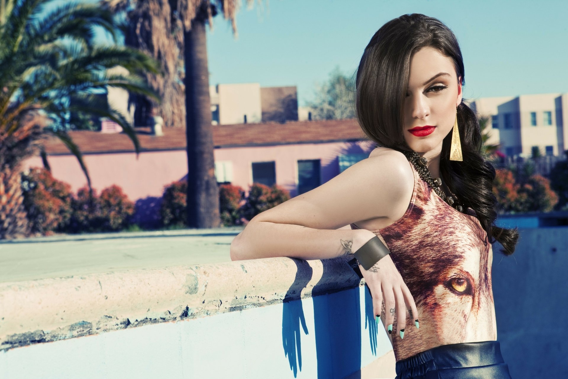 HD wallpaper of English singer Cher Lloyd with brunette hair and bold lipstick, posing outdoors against a sunny backdrop with palm trees and buildings.