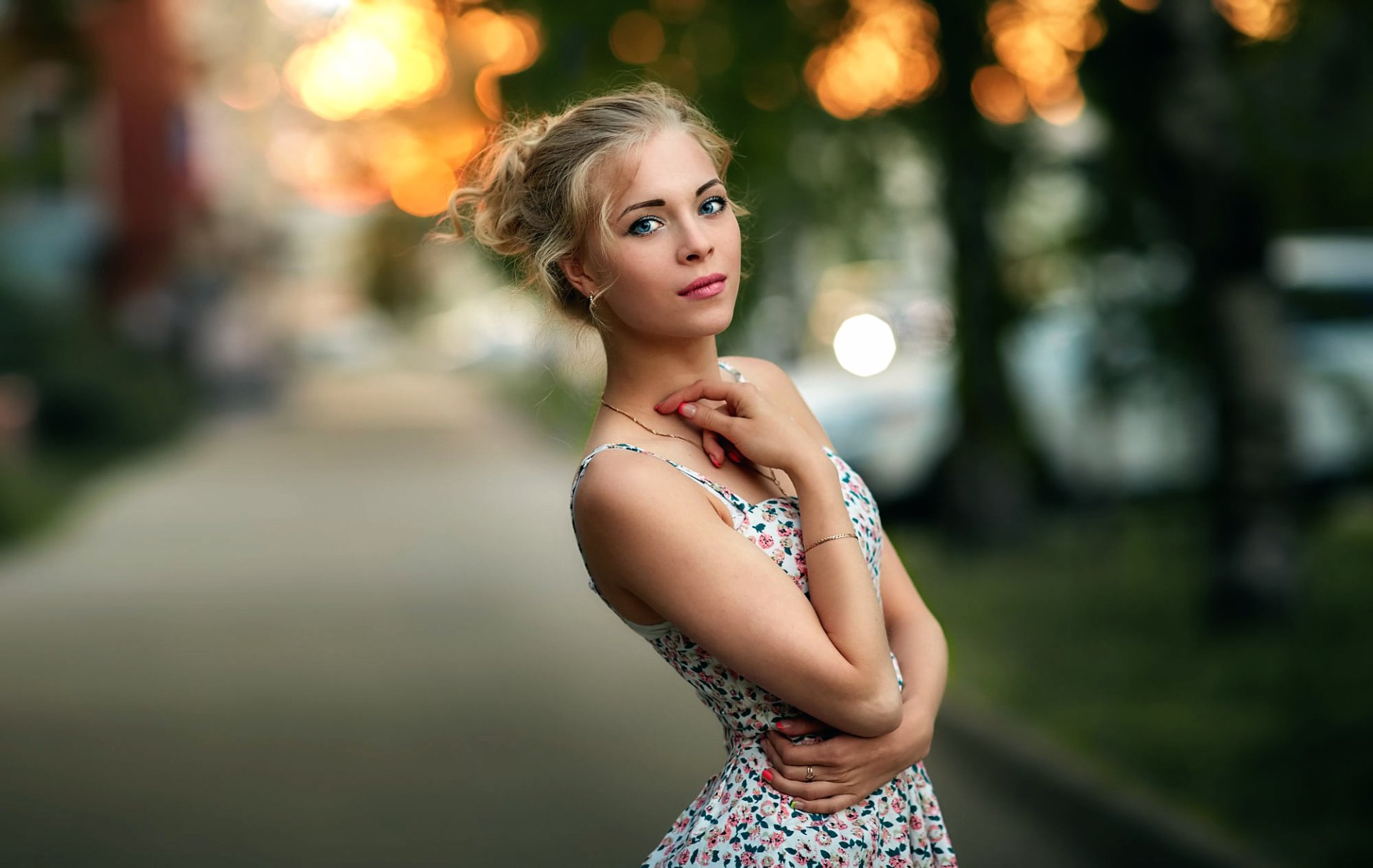 An outdoor HD wallpaper showing a blonde woman with blue eyes wearing a patterned dress, standing on a slightly blurred pathway with a bokeh background.