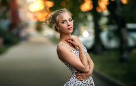 An outdoor HD wallpaper showing a blonde woman with blue eyes wearing a patterned dress, standing on a slightly blurred pathway with a bokeh background.