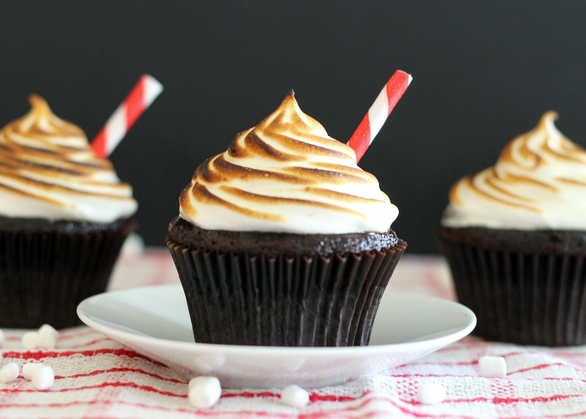 HD desktop wallpaper of three chocolate cupcakes topped with swirled cream and red striped straws, set against a dark background.