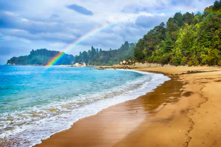 HD desktop wallpaper of a New Zealand beach featuring golden sand, lush coastline, and a vibrant rainbow arching over the serene ocean under a cloudy sky.