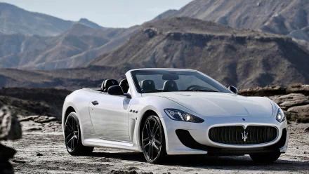 White Maserati GranTurismo convertible grand tourer parked on a rocky terrain with mountainous backdrop, shown in a high-definition PC desktop wallpaper.