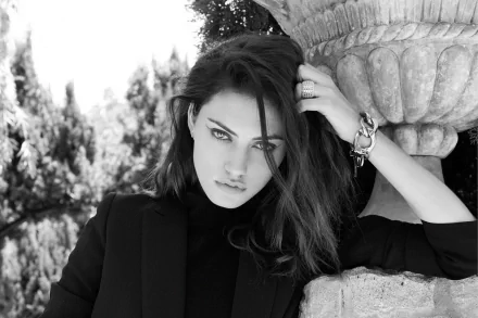 Black and white HD portrait of Australian actress and model Phoebe Tonkin leaning against a stone structure, her brunette hair framing a striking, confident expression.