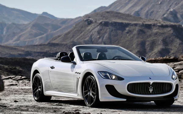 White Maserati GranTurismo convertible grand tourer parked on a rocky terrain with mountainous backdrop, shown in a high-definition PC desktop wallpaper.