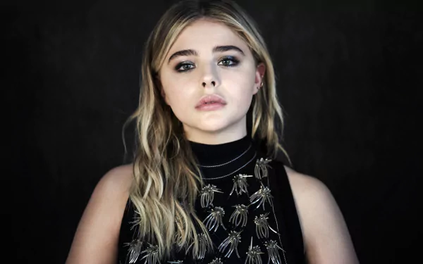 Blonde American actress Chloë Grace Moretz poses confidently, showcasing her striking green eyes. This HD wallpaper features her in an elegant outfit against a dark backdrop.