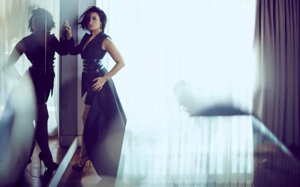 American singer Demi Lovato, a brunette with brown eyes, stands in a black dress beside her reflection in a sleek, modern interior, captured in HD.