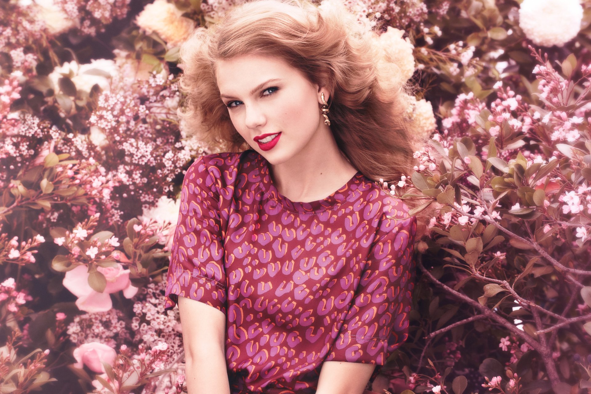 A vibrant image of a blonde American singer with blue eyes, smiling confidently, wearing lipstick and earrings, surrounded by blooming flowers. Ideal HD wallpaper for music fans.