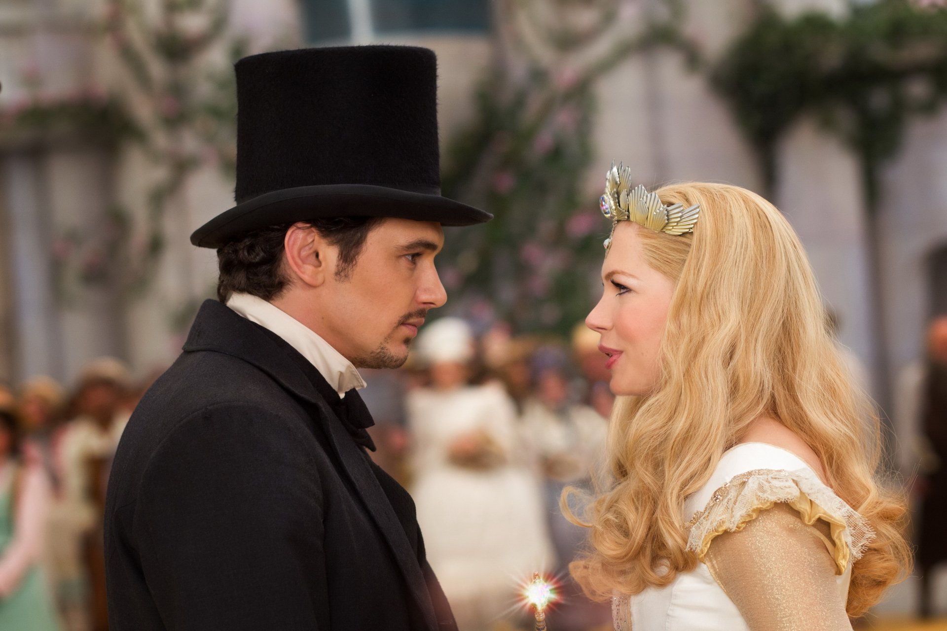 Two costumed characters from Oz the Great and Powerful — a man in a top hat and a blonde woman in white — face each other in an ornate ballroom. 2K Quad HD desktop wallpaper.