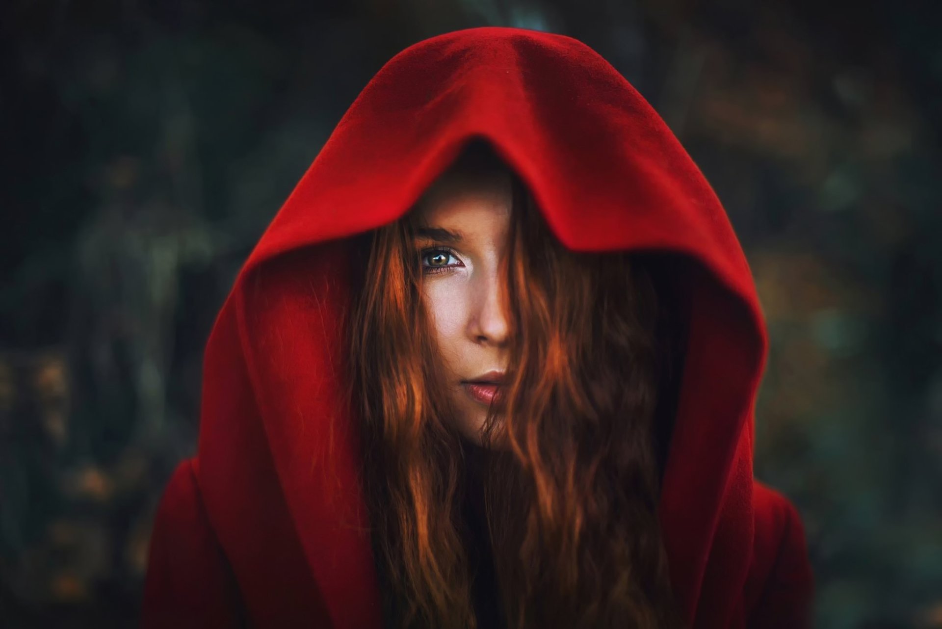 HD desktop wallpaper of a redhead woman model wearing a red hood, with intense blue eyes and a softly blurred dark background.