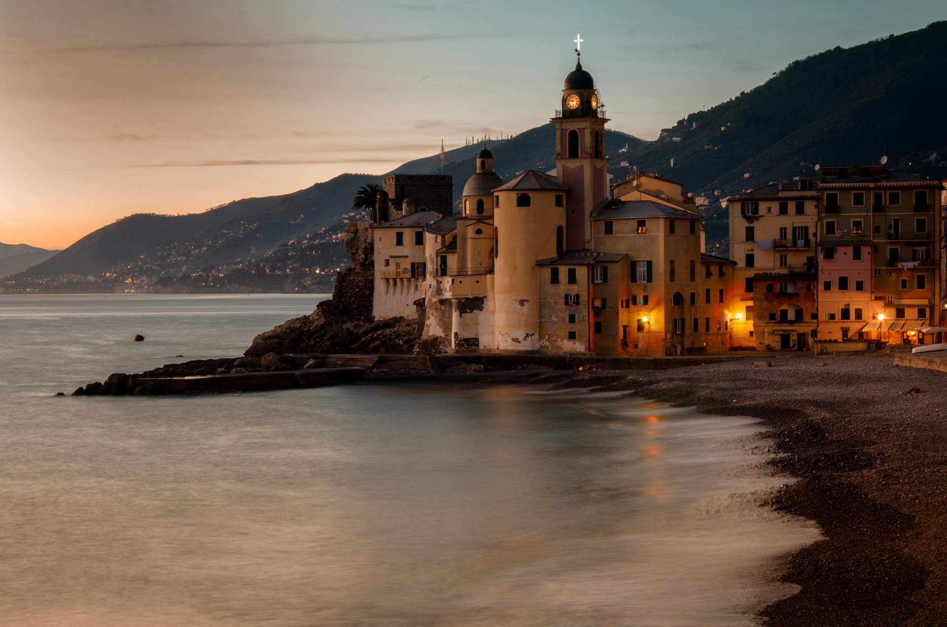 Evening view of the charming man-made town of Camogli on Italy’s coast, featuring illuminated buildings along the beach with mountains in the background.