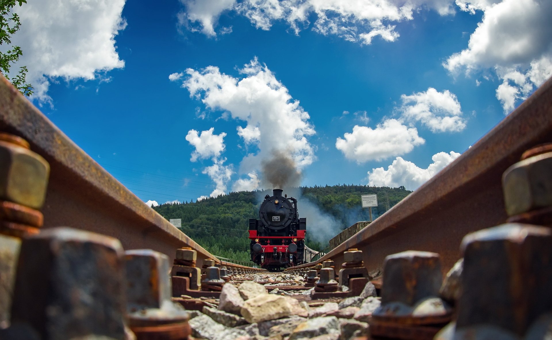 A steam locomotive approaches on railroad tracks under a bright blue sky, surrounded by lush greenery, creating a striking HD wallpaper for PC desktops.