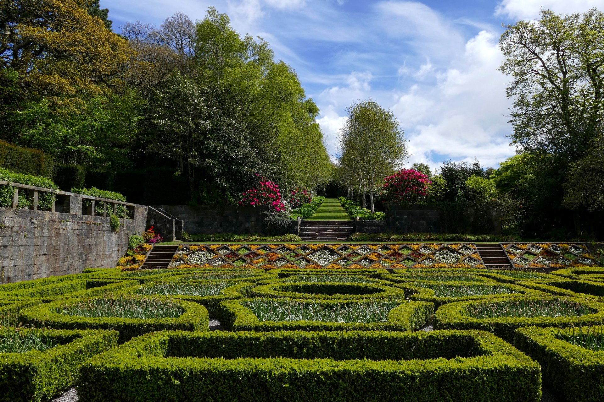 High-definition desktop wallpaper of a meticulously landscaped garden featuring hedge patterns, colorful flowers, and lush greenery under a partly cloudy sky.