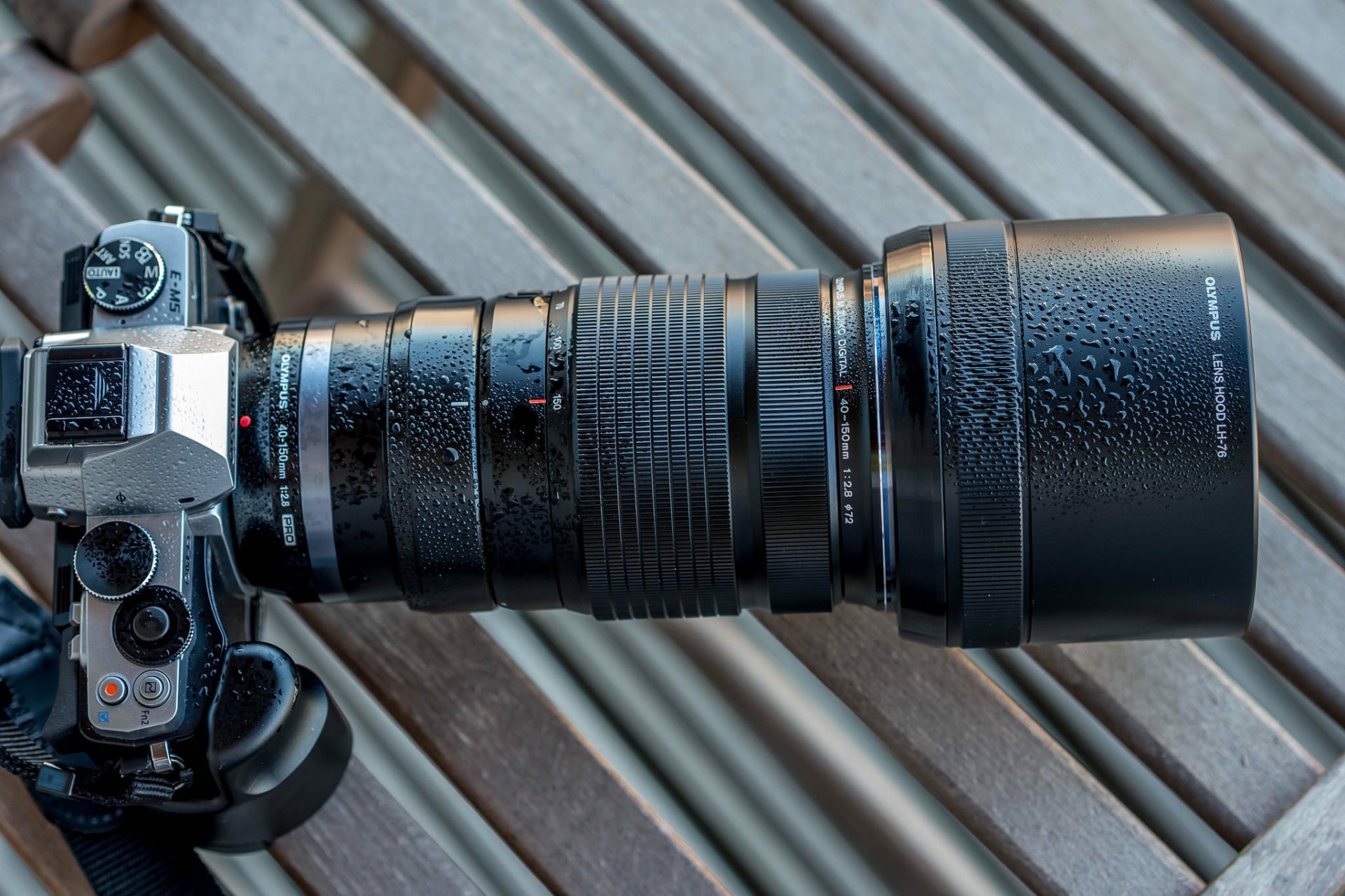 Close-up of an Olympus camera with a detailed man-made lens, showcasing water droplets on the surface, set against a wooden background in this HD desktop wallpaper.