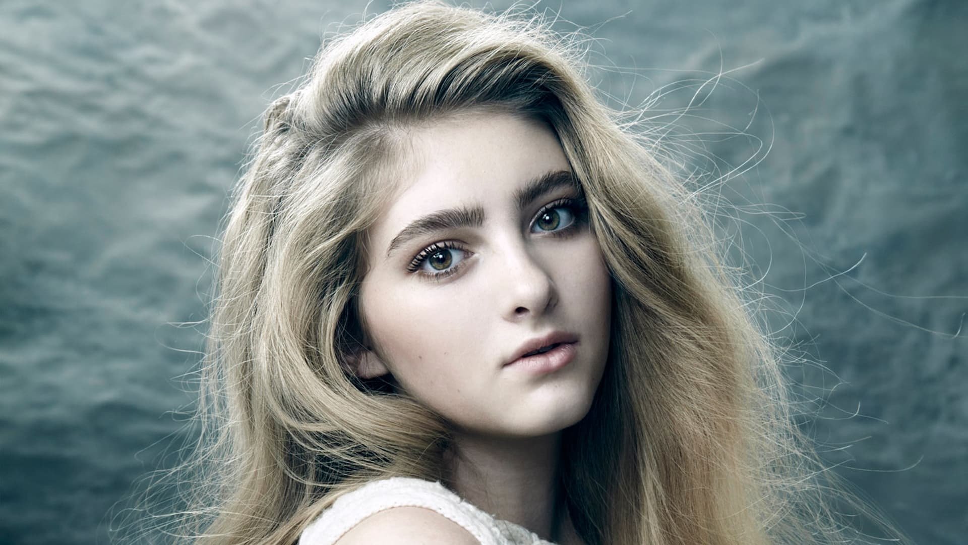 Close-up HD portrait of American actress Willow Shields with blonde hair and brown eyes, set against a textured gray background.