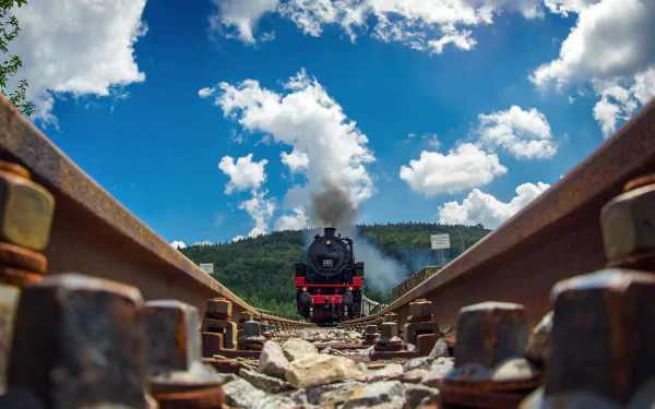 A steam locomotive approaches on railroad tracks under a bright blue sky, surrounded by lush greenery, creating a striking HD wallpaper for PC desktops.