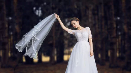 An Asian brunette model poses outdoors in a flowing wedding dress, holding a delicate veil against a blurred forest backdrop in this 4K Ultra HD desktop wallpaper.