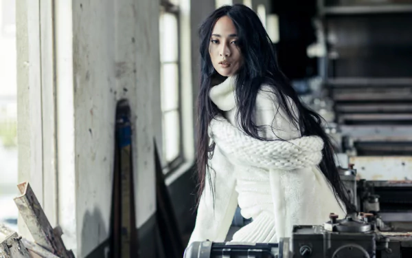 8K Ultra HD wallpaper of a blue-eyed woman model with long black hair in a white sweater and knit scarf, posed by a window in a weathered industrial interior.