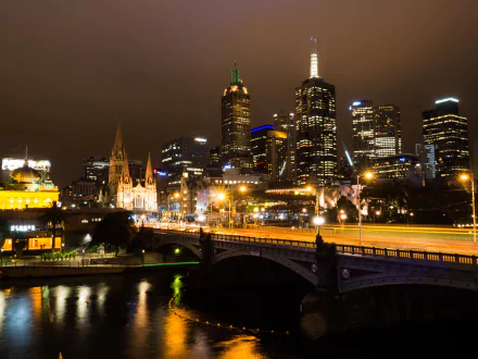 Nighttime view of Melbourne's skyline featuring illuminated skyscrapers, a bridge, and city lights reflecting on water in this 4K Ultra HD wallpaper of the Australian city.