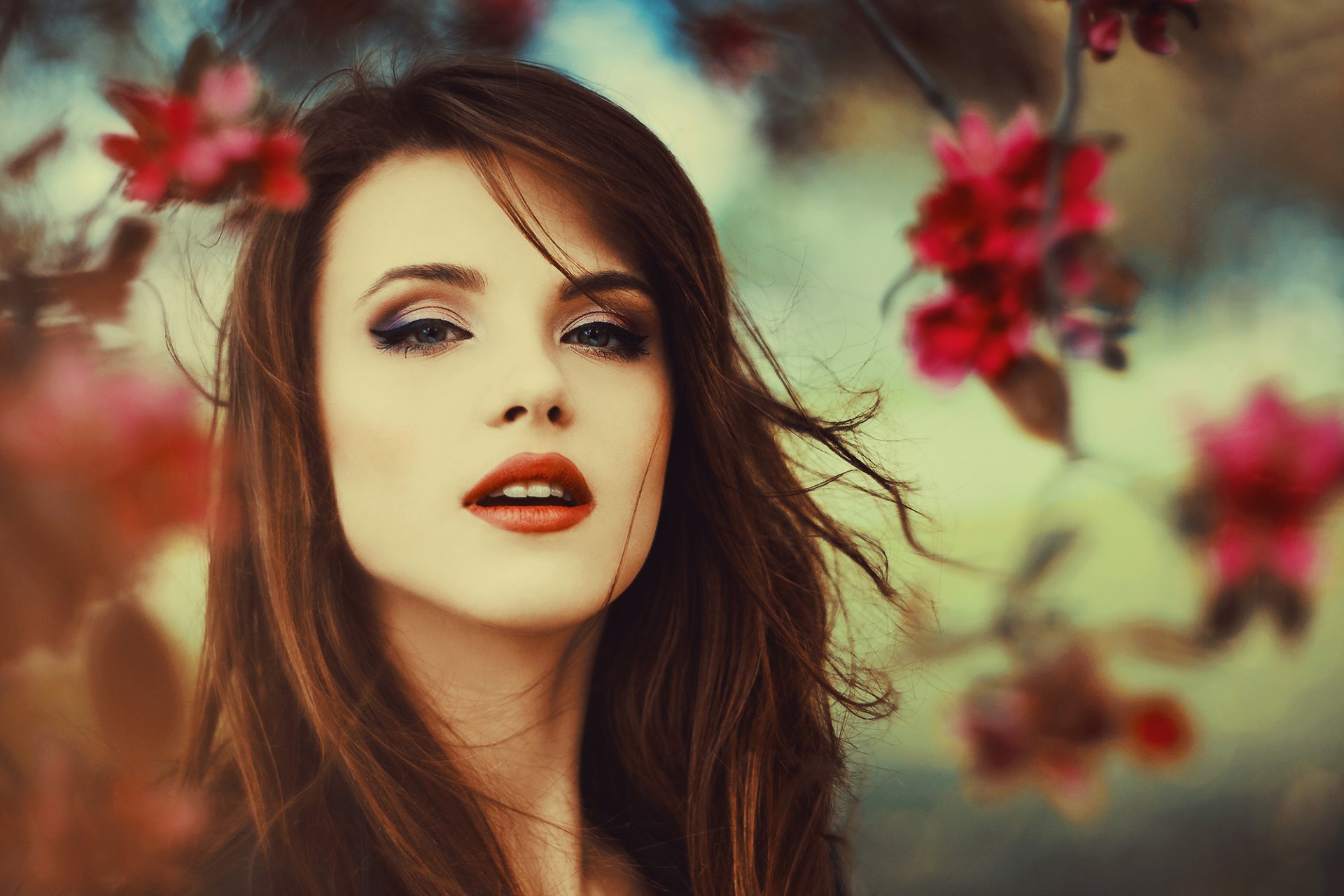 A brunette model with striking lipstick poses against a blurred backdrop of flowers, showcasing her expressive face in this HD desktop wallpaper.