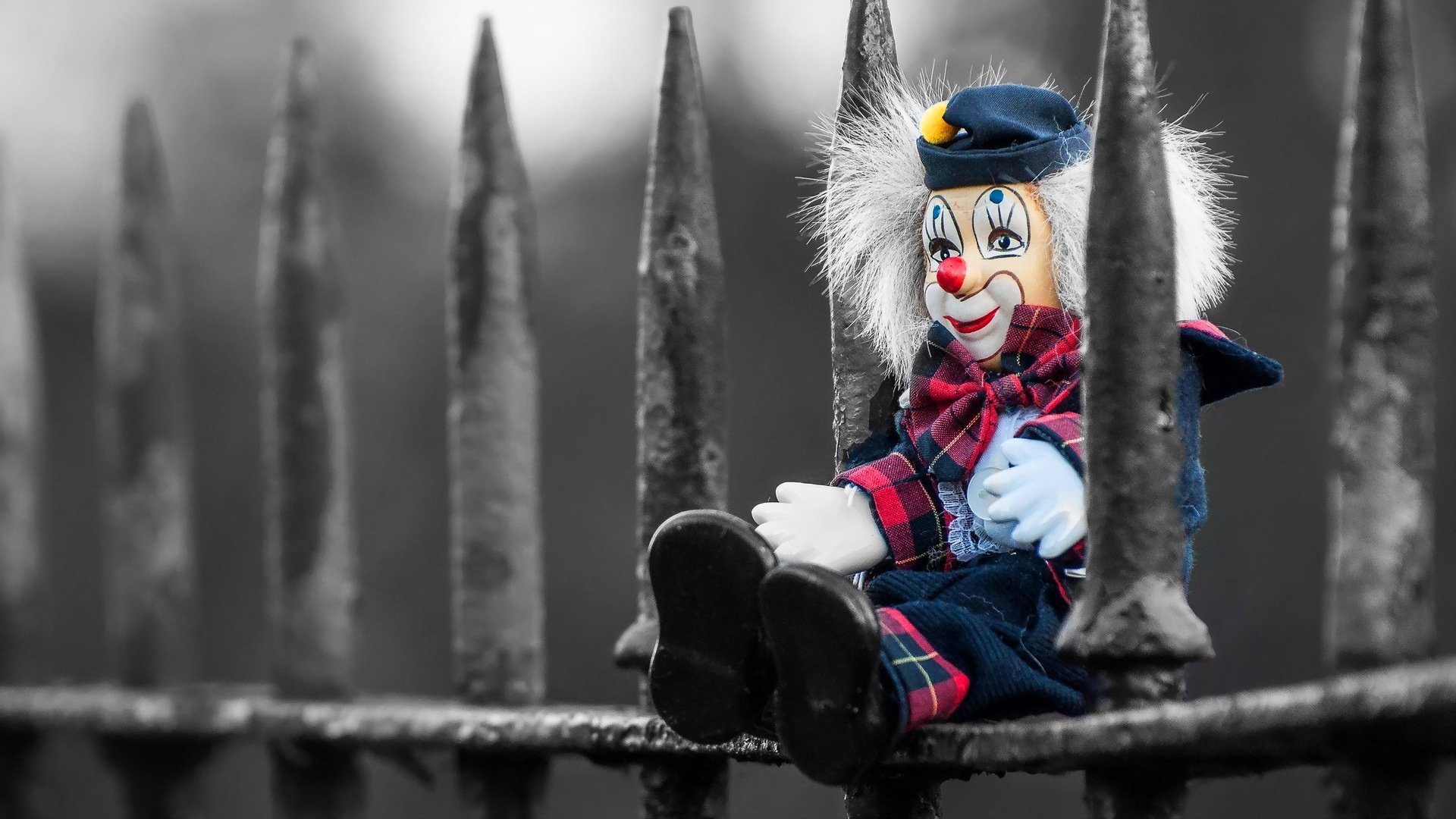 A colorful clown toy with a blue hat and red plaid shirt sits against a dark, spiked metal fence in this HD PC desktop wallpaper and background.