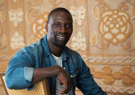 HD desktop wallpaper of French actor Omar Sy smiling against an ornate beige and gold patterned background.