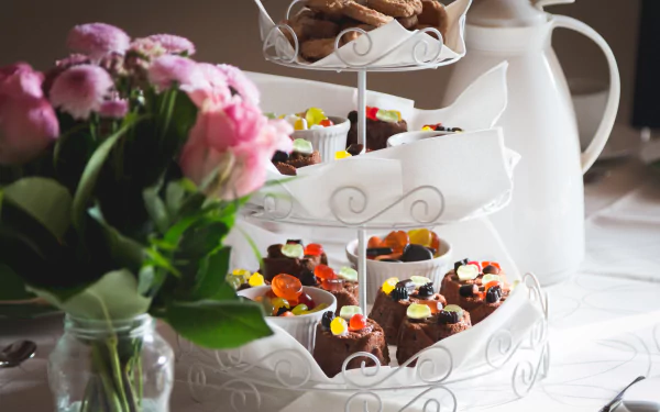 A 4K Ultra HD still life image of biscuits and colorful candy sweets arranged on a tiered stand with pink flowers in the foreground and a white pitcher in the background.