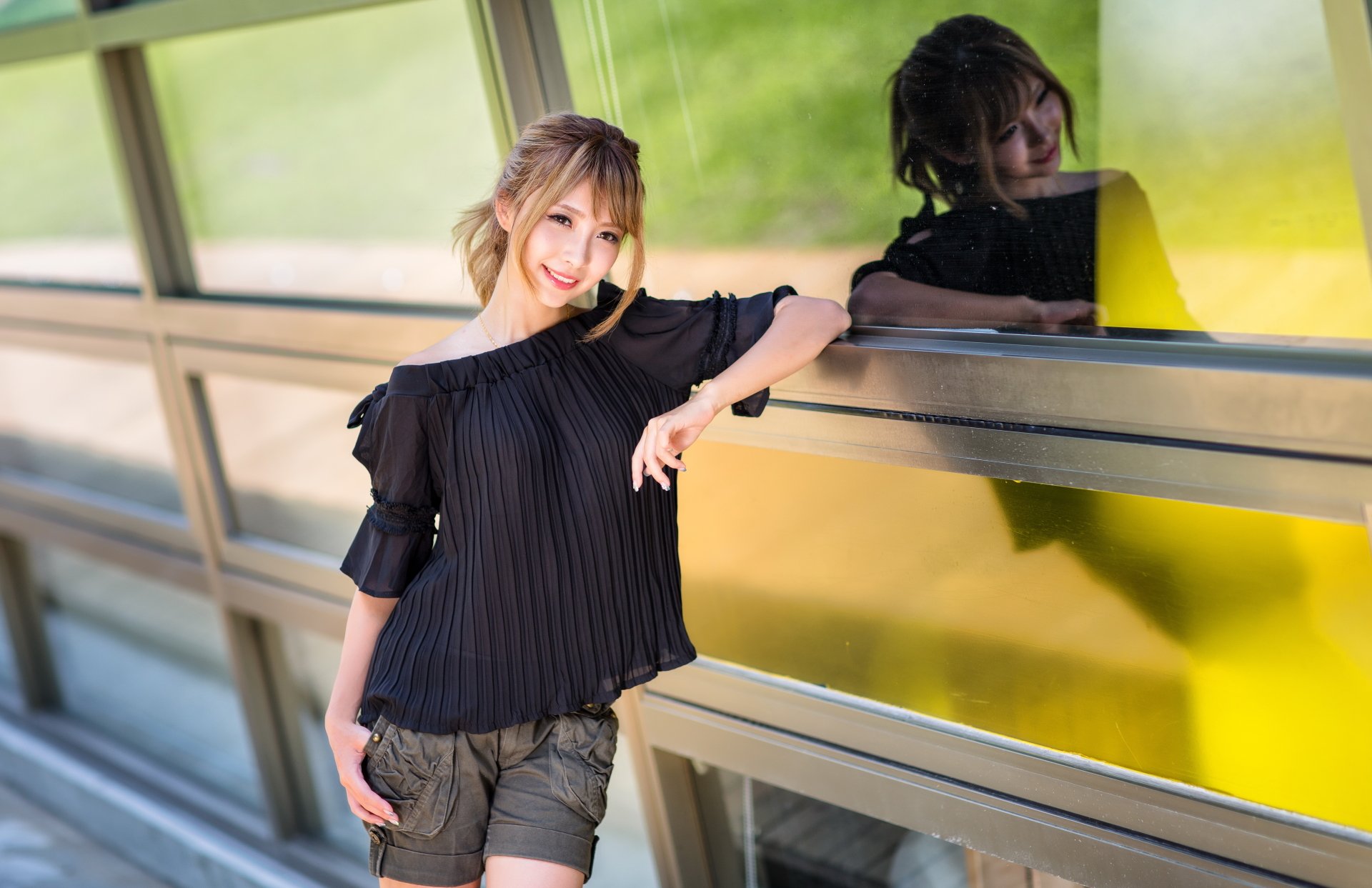 A brunette Asian woman stands smiling against a reflective surface, creating a mirrored image. She is dressed casually, and the background is an outdoor setting. This HD wallpaper exudes warmth and relaxation.