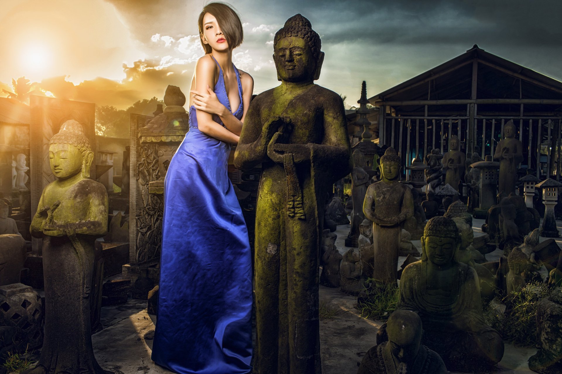 A woman with brunette hair in a blue dress poses beside a Buddha statue amidst a collection of stone figures, illuminated by a dramatic sunset, creating a serene atmosphere.