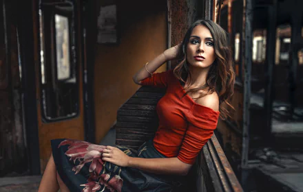 HD desktop wallpaper featuring a brunette woman with brown eyes, wearing a red top and floral skirt, posing thoughtfully in a rustic, dimly lit setting.