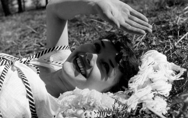 Black-and-white 2K Quad HD desktop wallpaper of a smiling vintage actress reclining on grass, shading her eyes with one hand, wearing a striped top with ruffled fabric beside her.