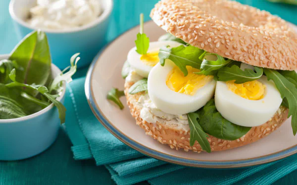 A close-up of a sesame bagel sandwich filled with sliced eggs and fresh greens, accompanied by a small bowl of salad. This vibrant 4K Ultra HD image captures a delicious meal.