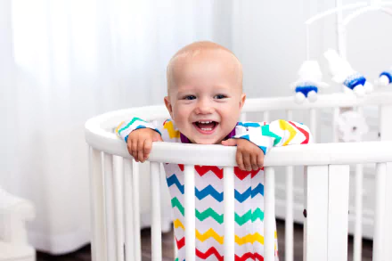 Smiling baby in a colorful chevron onesie leaning on a white crib rail — bright baby photography, 2K Quad HD PC desktop wallpaper/background.