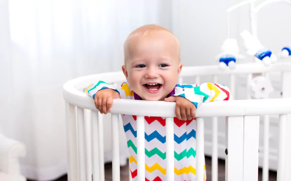 Smiling baby in a colorful chevron onesie leaning on a white crib rail — bright baby photography, 2K Quad HD PC desktop wallpaper/background.