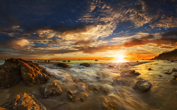 A stunning sunset over the ocean at Malibu, featuring vibrant sunbeams illuminating the horizon, with gentle waves lapping against rocky shores. 8K Ultra HD wallpaper.