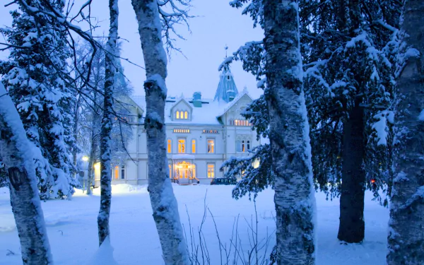Snow-covered trees frame Fjällnäs Castle in Sweden during winter, with warm lights glowing from its windows against a serene, snowy landscape.