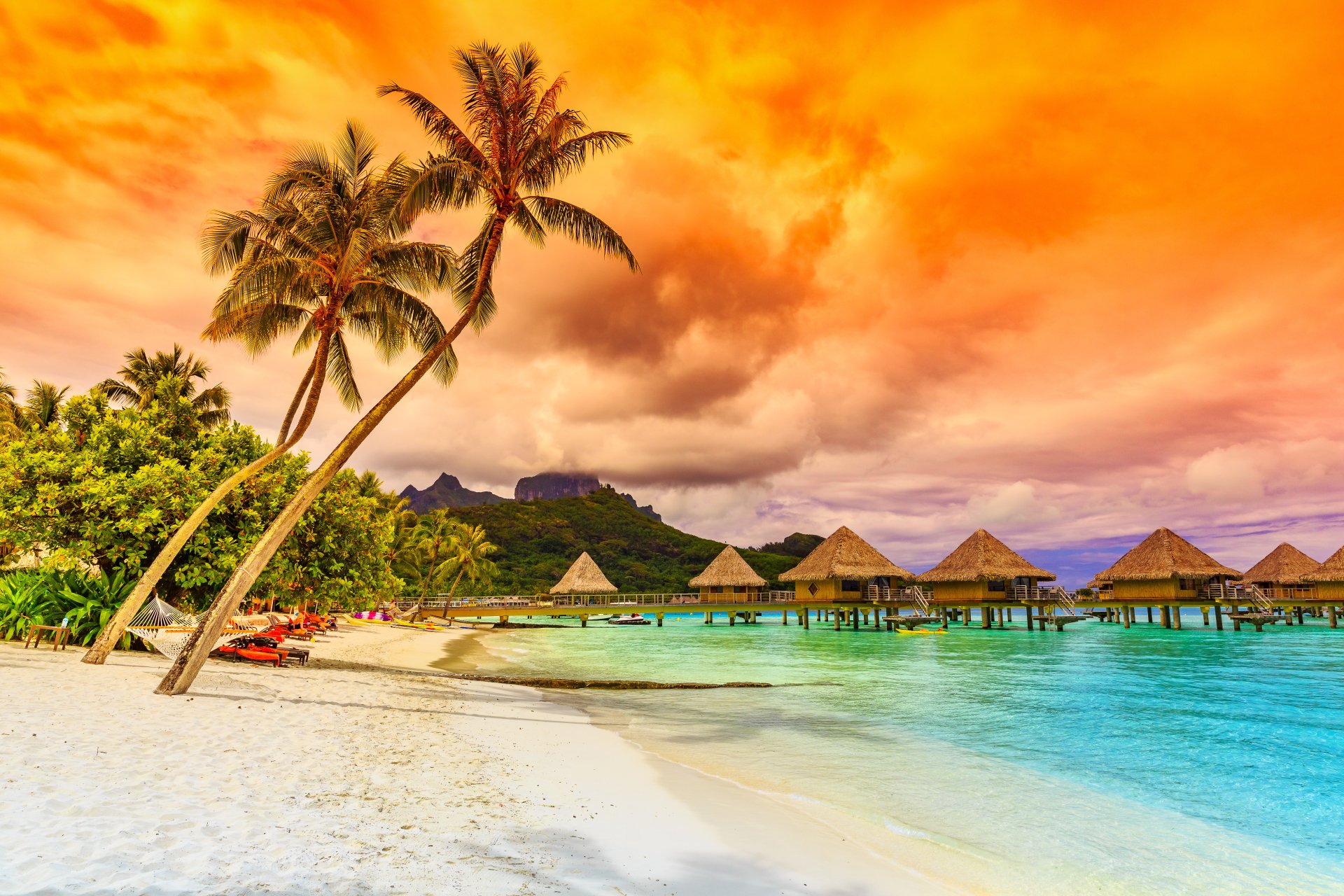 Tropical beach scene with palm trees, sandy shore, thatched huts over clear water, vibrant orange sky, captured in 4K Ultra HD for a stunning holiday backdrop.