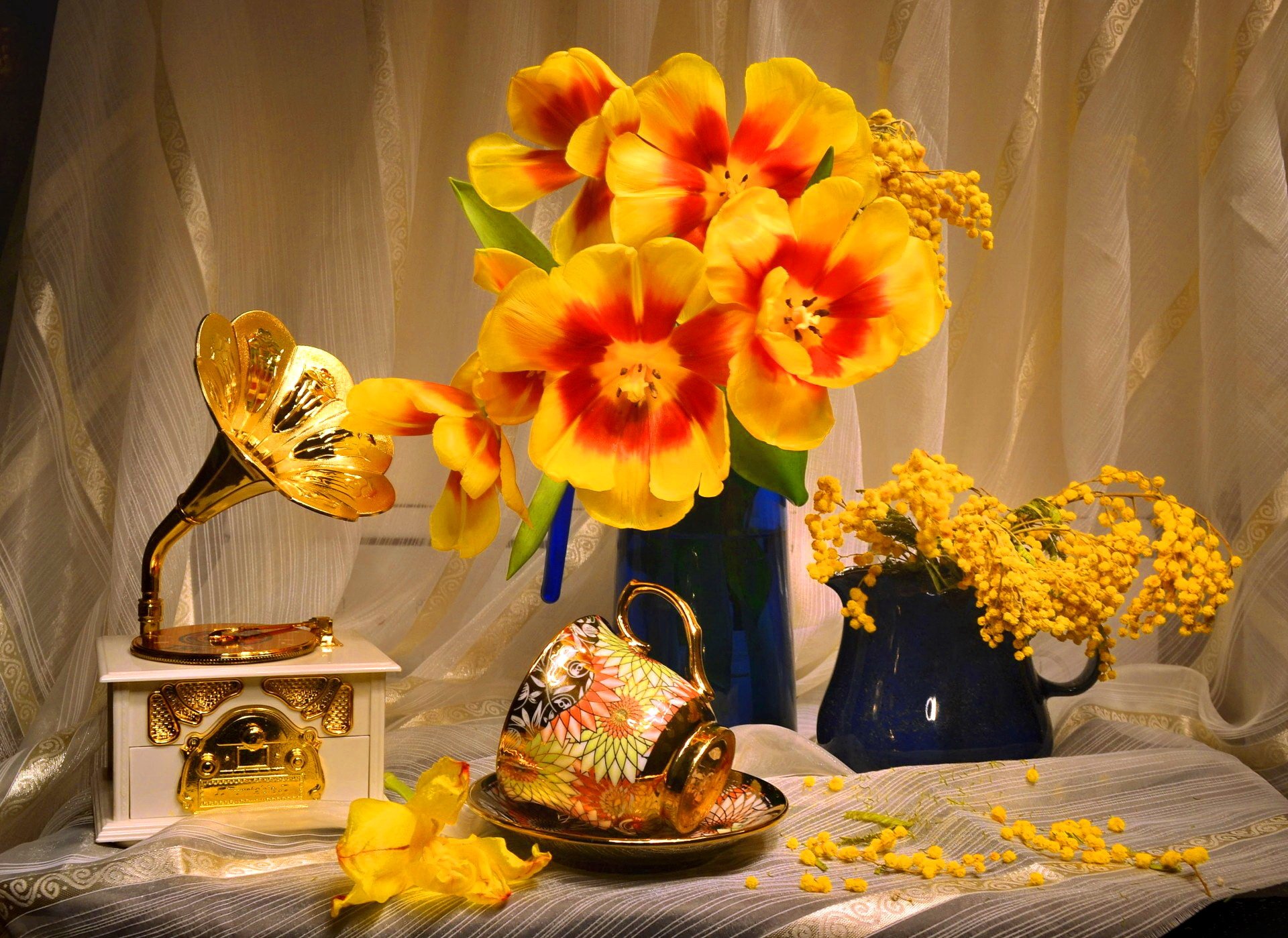 HD desktop wallpaper featuring a still life of vibrant yellow and orange tulips in a vase, a vintage gramophone, and a decorative cup arranged on a draped fabric background.