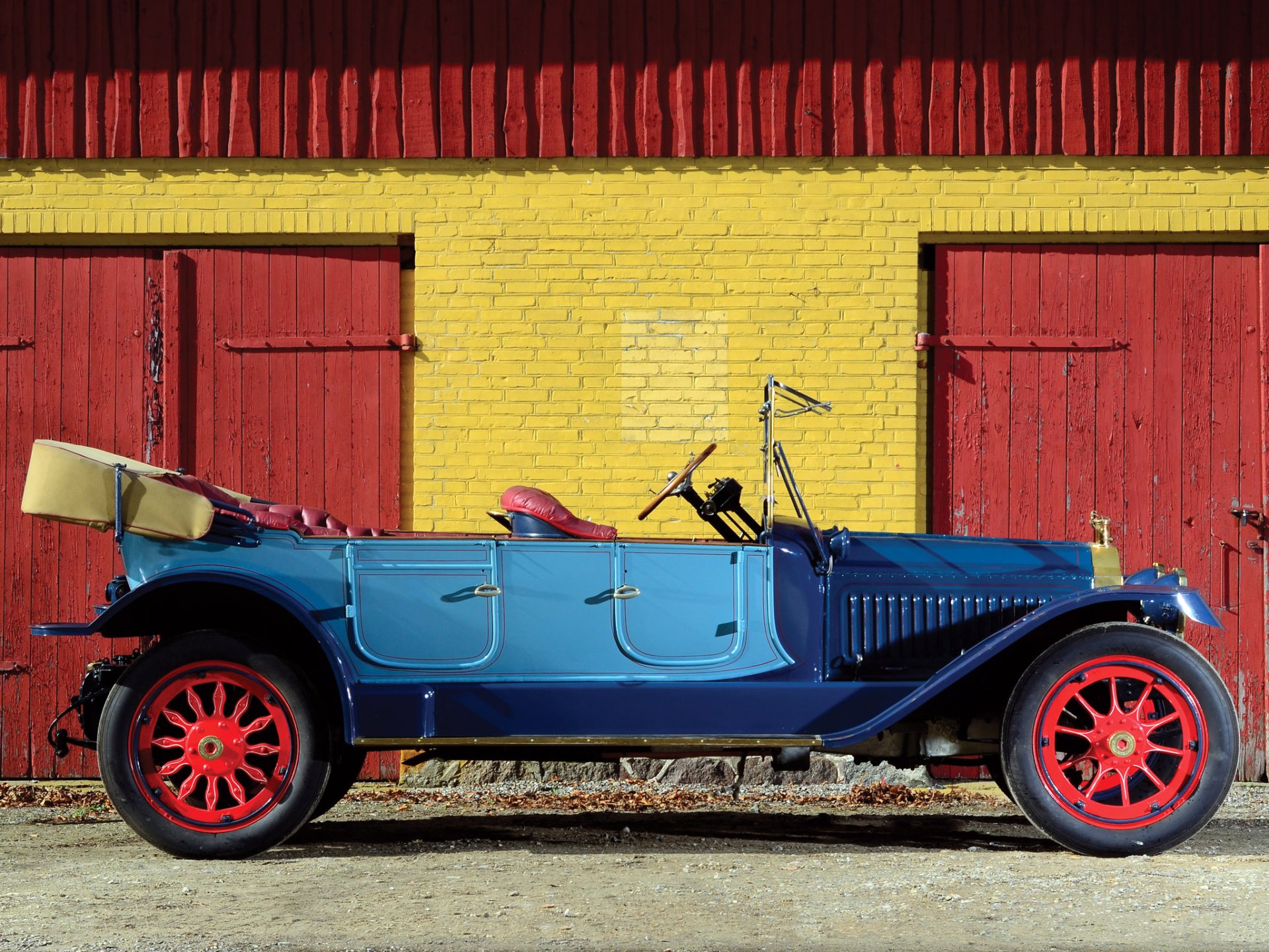 Side view of a blue 1914 Packard Six 7-passenger Touring vintage car with red wheels parked before a yellow brick wall and red doors — HD PC desktop wallpaper