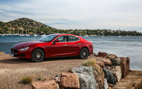 A red Maserati Ghibli car parked near rocky shore with calm water and hills in the background, presented as a 4K Ultra HD PC desktop wallpaper.