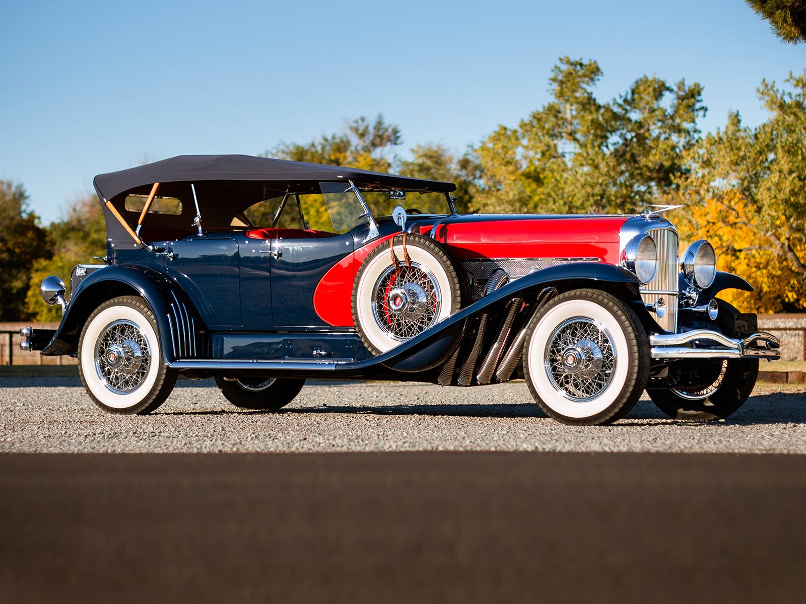 Vintage 1929 Duesenberg Model J Dual Cowl Phaeton with black and red body, whitewall tires, and convertible top, set against a scenic outdoor background.