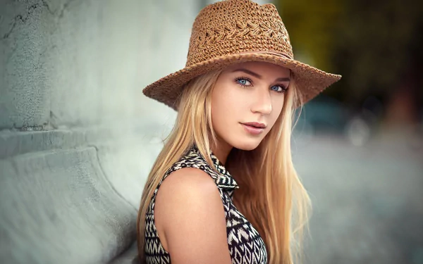 A blonde model with blue eyes and a stylish hat poses against a textured wall, showcasing her vibrant look. This HD image serves as an eye-catching desktop wallpaper.