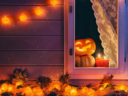 A cozy Halloween scene featuring a glowing jack-o'-lantern and candles by a window, surrounded by autumn leaves and festive lights, creating a warm night atmosphere.