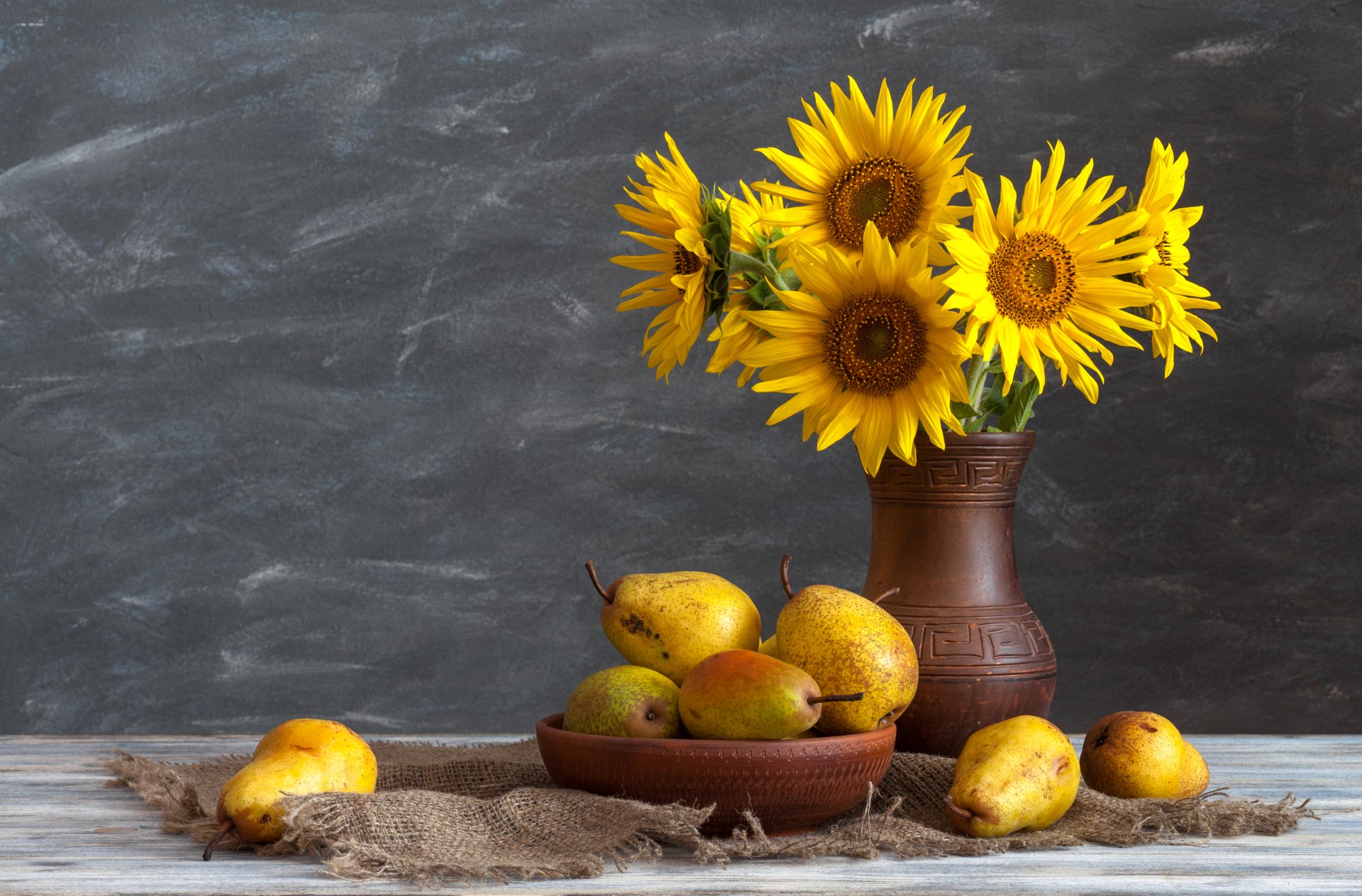 8K Ultra HD desktop wallpaper: still life of bright yellow sunflowers in a ceramic vase beside a bowl of pears on a rustic table.