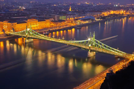 Night view of a brightly lit man-made bridge spanning the Danube River in Budapest, Hungary, with city lights reflecting on the water.