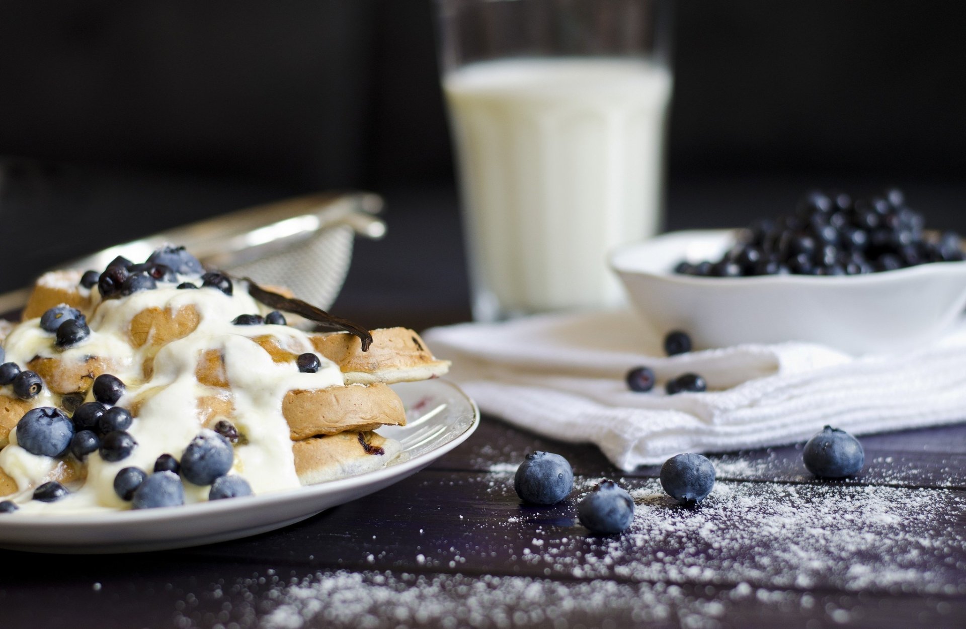 HD desktop wallpaper featuring a stack of blueberry waffles topped with cream, fresh blueberries, and a glass of milk in a cozy breakfast setting.