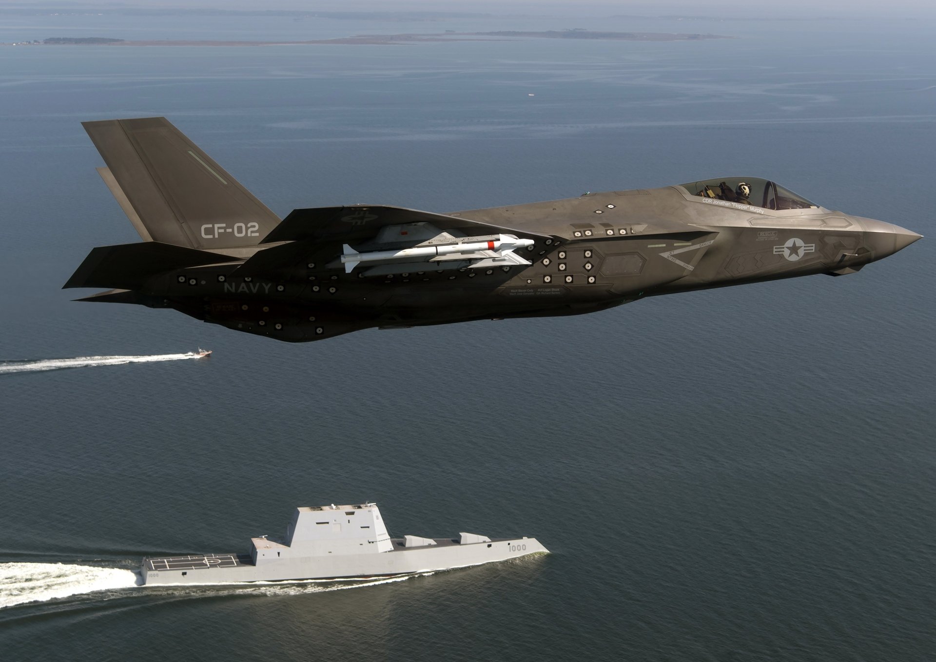 An F-35 Lightning II jet flies over the USS Zumwalt (DDG-1000) warship, showcasing advanced military technology against a serene ocean backdrop.