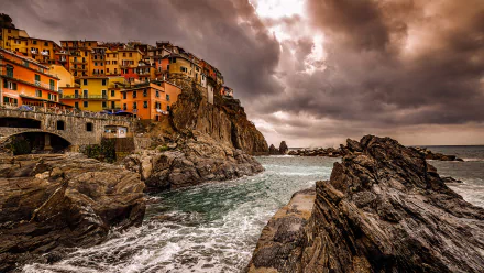 HD desktop wallpaper of Manarola in Italy's Cinque Terre, showcasing colorful houses on rocky cliffs by the ocean under dramatic cloudy skies.