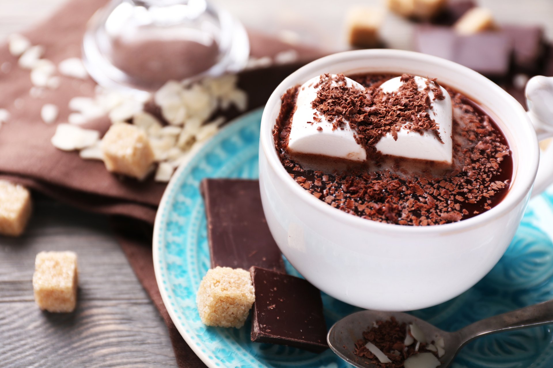 Close-up of a cup of hot chocolate topped with marshmallows and grated chocolate, surrounded by sugar cubes and chocolate pieces on a blue plate, in 4K Ultra HD quality.