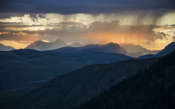 A stunning Montana sunset illuminates the clouds over the mountains, showcasing the dramatic landscape of Glacier National Park in a breathtaking wilderness scene.