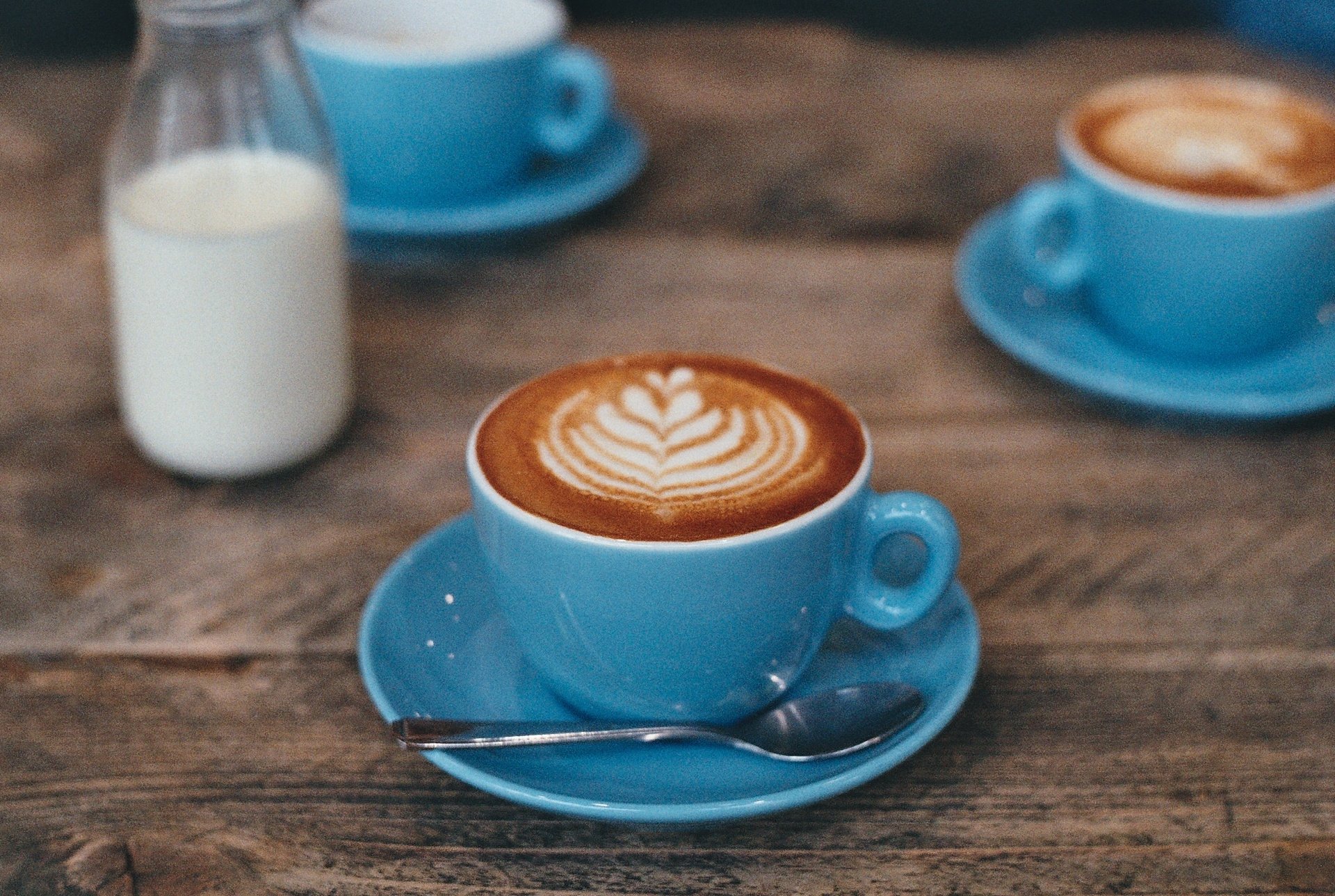 HD desktop wallpaper featuring a close-up of a blue cup filled with coffee topped with latte art, accompanied by a small bottle of milk on a rustic wooden table.